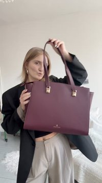 Bordo spalvos odinė rankinė su dviem rankenomis, auksinės spalvos metalinėmis detalėmis ir formos aiškumu. Priekyje yra prekės ženklo logotipas.