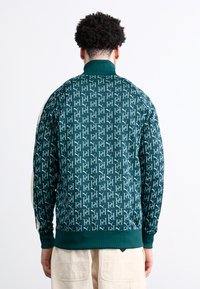 Veste verte à motifs avec un motif de logo répété, col et poignets côtelés, et une coupe ajustée. Fabriquée en tissu texturé.