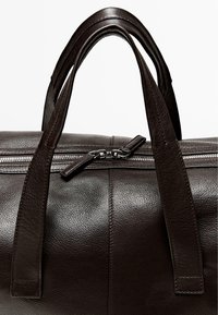 Dunkelbraune Ledertasche mit glatter Struktur, ausgestattet mit zwei stabilen Griffen und einem silbernen Reißverschluss. Auffällige Nähte betonen das Design.