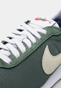 Zöld vászon sportcipő fehér fűzőkkel, bézs Nike swoosh logóval és párnázott nyelvvel. Texturált felsőrész és fehér gumitalp.
