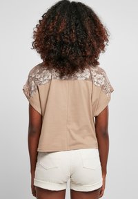 Kortærmet bluse i beige med ryg i blonder og rund halsudskæring. Sat sammen med hvide shorts med opsmøg og krøllet hår med tekstur.