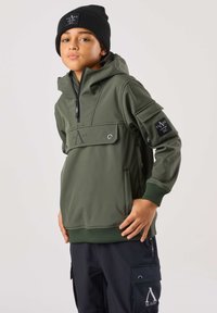 Grøn hættejakke med en forlom, lynlås og logodetalje. Bærer med en sort hue og mørke cargobukser med sidelommer.