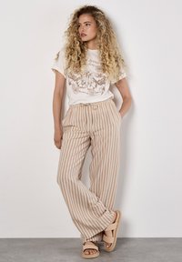 Pantalons larges beige clair à rayures verticales brunes, associés à un t-shirt blanc avec un motif de papillon. Modèle portant des sandales.