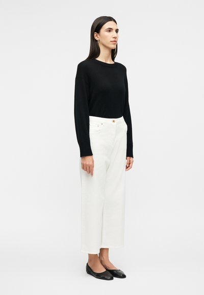 Γυναίκα που στέκεται στραμμένη προς τα δεξιά, ντυμένη με μαύρη μπλούζα μακρυμάνικη, άσπρο παντελόνι με φαρδιά μπατζάκια και μαύρες μπαλαρίνες, μπροστά σε απλό φωτεινό φόντο.