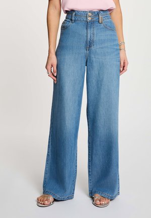 Vaqueros azules de talle alto y pierna ancha con doble botón y cremallera, combinados con una blusa rosa claro y sandalias plateadas.