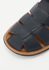 Jūras zila ādas sandele ar atvērtu purngalu, kurai ir pinumi un kontrastējošas šuves. Zole ir dabīgs brūns tonis ar strukturētu apdari.