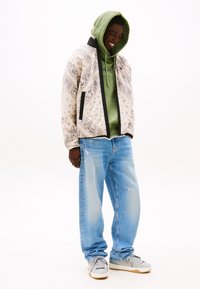 Men's outfit featuring a patterned cream jacket with black accents, green hoodie, light blue distressed jeans, and gray sneakers.