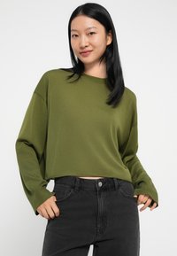 Fiatal nő hosszú fekete hajjal, olíva zöld, hosszú ujjú cropped toppot és magas derekú fekete farmernadrágot visel, kezével a zsebei közel áll.