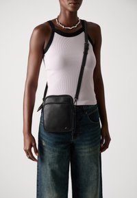 Borsa a tracolla nera in pelle con superficie testurizzata, forma rettangolare, tracolla regolabile e chiusura con zip, indossata sopra un top in lilac.