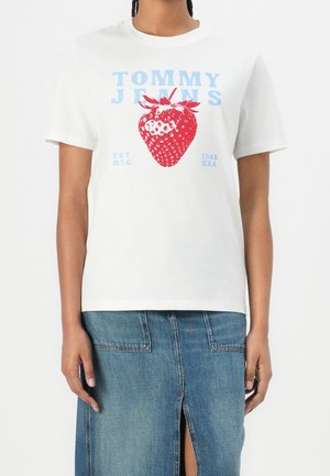Biała koszulka z niebieskim napisem "Tommy Jeans", dużą czerwoną grafiką truskawki, noszona z niebieską spódnicą dżinsową z przednim rozcięciem i naszywkami na kieszeniach.