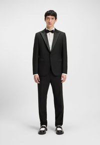Mees seisab, kandes musta smokingut satiinist kraede, valget triiksärki, musta kikilipsu, musti pükse ja mustvalgeid kingi.