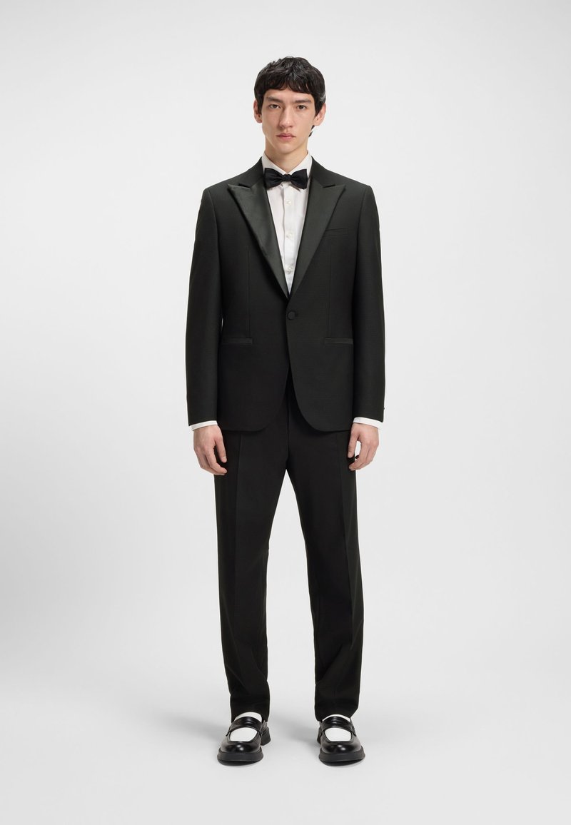 Mees seisab, kandes musta smokingut satiinist kraede, valget triiksärki, musta kikilipsu, musti pükse ja mustvalgeid kingi.