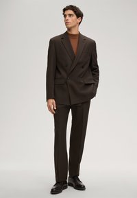 Doppelreihiger dunkelbrauner Anzug mit gekerbtem Revers, kombiniert mit einem taillierten braunen Rollkragenpullover und dunklen Schuhen, mit schmalem Schnitt und maßgeschneiderter Passform.