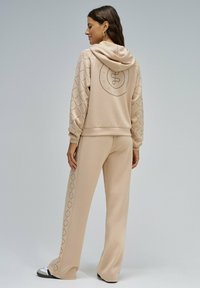 Sudadera con capucha beige y pantalones de chándal a juego, con un gráfico en la parte posterior y patrones geométricos en las mangas y los lados de los pantalones.