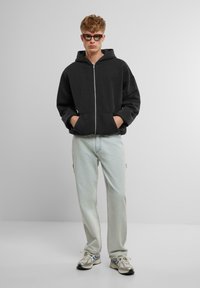 Črna zip-up kapuca s prostornim kapuco, sproščenega kroja, v kombinaciji z svetlo modrimi kavbojkami in sivimi supergami, postavljena proti enotni podlagi.