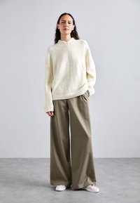 Crème kleurige gebreide trui met V-hals en vallende schouders, gecombineerd met losse, olijfgroene broek met wijde pijpen en witte sneakers.