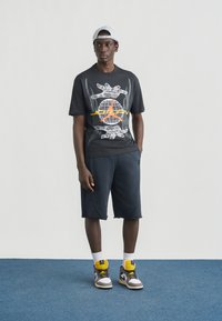 Μαύρο μπλουζάκι με γραφικό σχέδιο, γκρι σορτς και πολύχρωμα αθλητικά μποτάκια. Το μοντέλο φοράει ανοιχτό καπέλο και στέκεται σε μπλε δάπεδο.