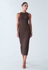 Ruskea ribbed midi-mekko, jossa on tiukka siluetti, pyöreä pääntie ja hienovarainen rakenne. Yhdistettynä remmisandaaleihin ja kultaisiin asusteisiin.