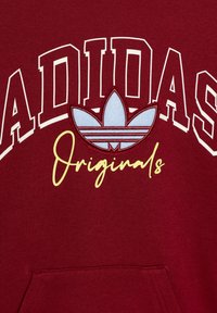 Vinröd sweatshirt med vit text "ADIDAS" och ljusblå Trefoil-logga. "Originals" i gul kursiv stil under loggan. Material i bomullsblandning.