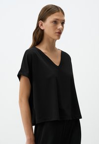 Top nero a maniche corte con scollatura a V, realizzato in tessuto morbido, caratterizzato da una vestibilità ampia e un orlo leggermente abbassato, con cuciture pulite e minime e senza motivi.