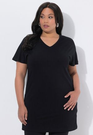 Kvinna med långt mörkt hår, bär pärlörhängen och en svart V-ringad klänning med korta ärmar, stående mot en vit bakgrund.