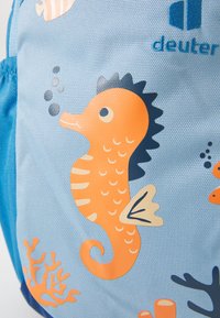 Zaino blu con una superficie testurizzata, arricchito da illustrazioni di cavallucci marini e corallo arancione e crema, completato dal logo del marchio in blu.