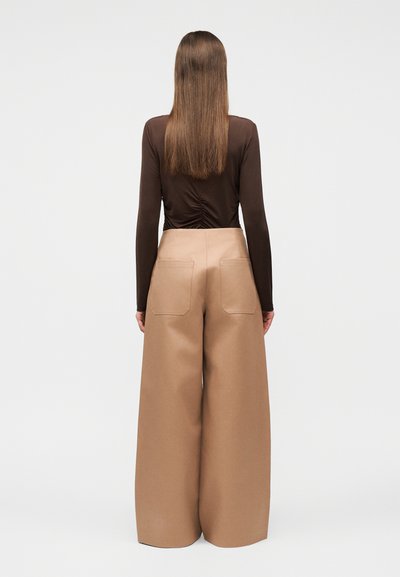 Άτομο με μακριά ίσια μαλλιά, που φοράει καφέ μπλούζα με μακριά μανίκια και μπεζ παντελόνι φαρδιάς γραμμής, στέκεται με την πλάτη στο πρόσωπο σε απλό φόντο.