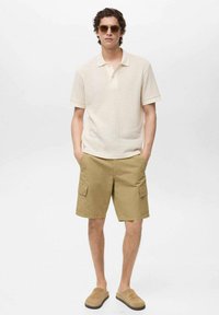 Polo shirt beige chiaro testurizzato con maniche corte, abbinato a pantaloni cargo khaki con tasche laterali. Indossato con scarpe slip-on color sabbia e occhiali da sole.