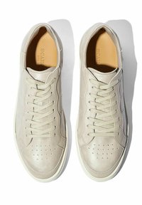 Bež kožne tenisice s glatkom teksturom, okruglim prstom, ukrasnim perforacijama i ravnim vezicama. Bijela guma, minimalistički dizajn.