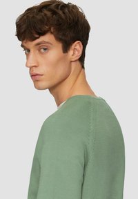 Pull en tricot vert avec un col rond, des poignets côtelés et une texture lisse. Détails de couture visibles au niveau des épaules.