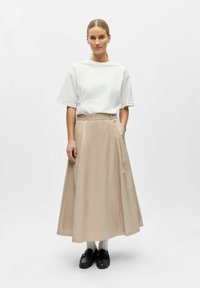 Femme debout portant une chemise blanche à manches courtes, une jupe beige longueur cheville, des chaussettes blanches et des mocassins noirs, sur un fond uni.