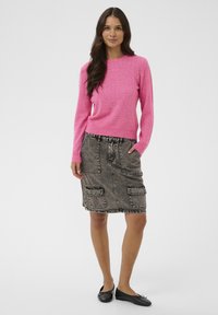 Femme debout portant un pull en tricot rose vif, une jupe en denim grise avec de grandes poches, et des ballerines noires sur un fond uni.