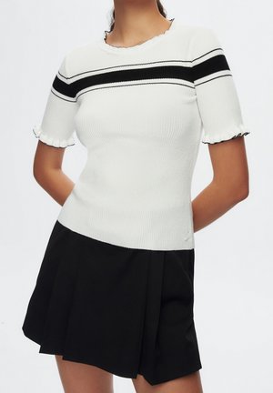 Vrouw draagt een wit geribd shirt met korte mouwen, zwarte strepen en gerimpelde randen, gecombineerd met een zwarte geplooide rok.