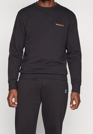 Homme portant un sweat Timberland noir et un pantalon noir assorti avec le logo Timberland sur la poitrine et la poche du pantalon.