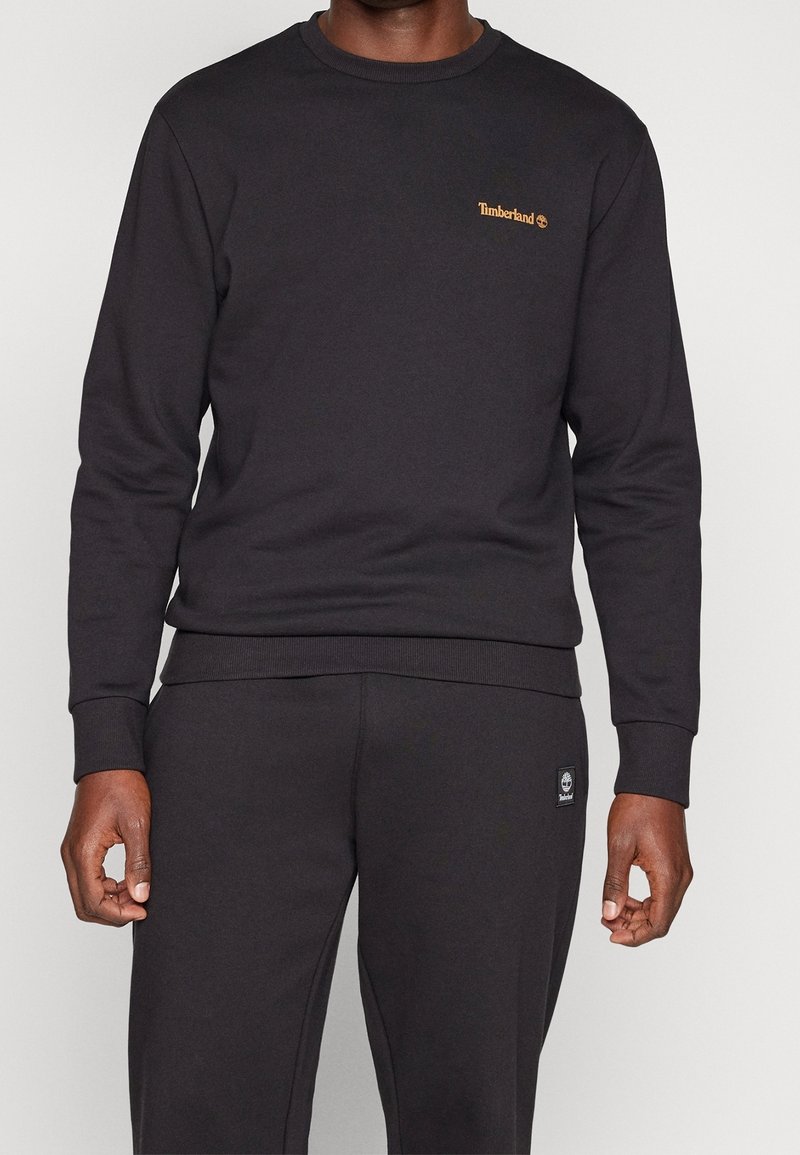 Homme portant un sweat Timberland noir et un pantalon noir assorti avec le logo Timberland sur la poitrine et la poche du pantalon.