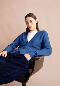 Junge Frau mit glattem braunen Haar, die eine blaue Strickjacke und dunkelblaue Cordhosen trägt, sitzt in einem braunen Stuhl vor einem beigen Hintergrund.