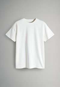 Fehér, rövid ujjú, kerek nyakú póló lóga egy szürke fal előtt, sima, egyszínű anyagot és laza szabást mutatva.