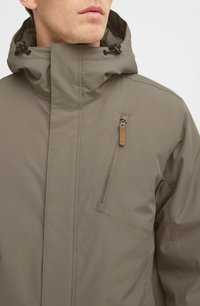 Chaqueta marrón, impermeable, con un alto cuello, cordones ajustables, bolsillo lateral con cremallera y textura suave y mate.