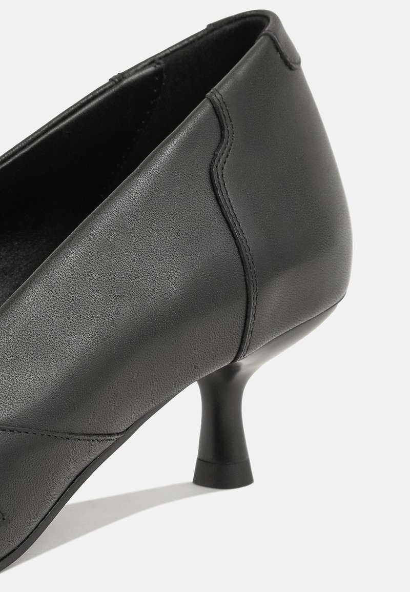 Zwarte leren schoen met een gebogen ontwerp, lage uitlopende hak, zachte textuur en minimalistische stiksels.
