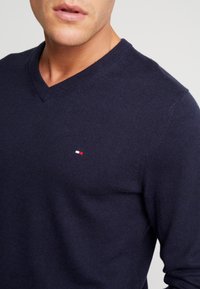 Granatowy sweter w serek wykonany z miękkiego materiału dzianinowego, z małym logo w czerwono-białych kolorach na lewej piersi oraz wygodnym krojem.