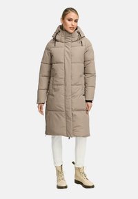 Kvinna som bär en knälång beige vadderad jacka med huva, vita byxor och beige snörade stövlar, stående mot en enkel ljus bakgrund.