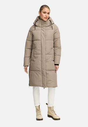 Femme portant un manteau rembourré beige à hauteur du genou avec capuchon, un pantalon blanc et des bottes beiges à lacets, se tenant devant un fond clair et uni.
