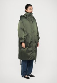 Manteau rembourré vert olive avec un col haut, une capuche et des poches latérales. Présente une texture lisse et brillante ainsi que des cordons de serrage ajustables à l'ourlet.