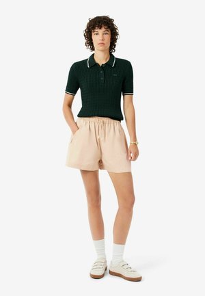 Donna in piedi, indossa una polo in maglia verde scuro a maniche corte, pantaloncini beige con coulisse, calzini bianchi e scarpe da ginnastica bianche, con le mani nelle tasche.