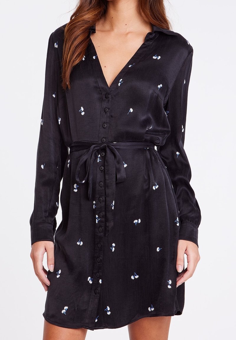 Femme portant une robe noire boutonnée avec de petits imprimés floraux blancs et bleus et une ceinture nouée à la taille, montrée du cou jusqu'à mi-cuisse.