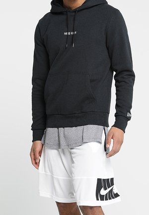 Sort hættetrøje med frontlomme og snore, lagdelt over en grå skjorte, kombineret med hvide meshshorts med et sort Nike-logo.