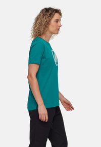 Teal bomulls t-shirt med rund hals, kort ärm och sidoklämmor; har en vit grafik på framsidan. Svarta byxor bärs nedan.