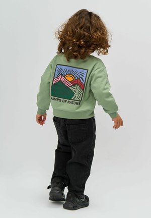 Felpa verde con grafica di montagna e testo "RAMPS OF NATURE", abbinata a jeans neri e sneakers nere, vista da dietro.