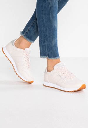 Persoon die lichtroze sportschoenen draagt met rubberen zolen en metallic zilverkleurige hielaccenten, gecombineerd met versleten blauwe spijkerbroeken.