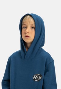 Modrá bavlnená mikina s veľkou kapucňou, s okrúhlym logom na hrudi. Mäkkučká textúra a uvoľnený strih.